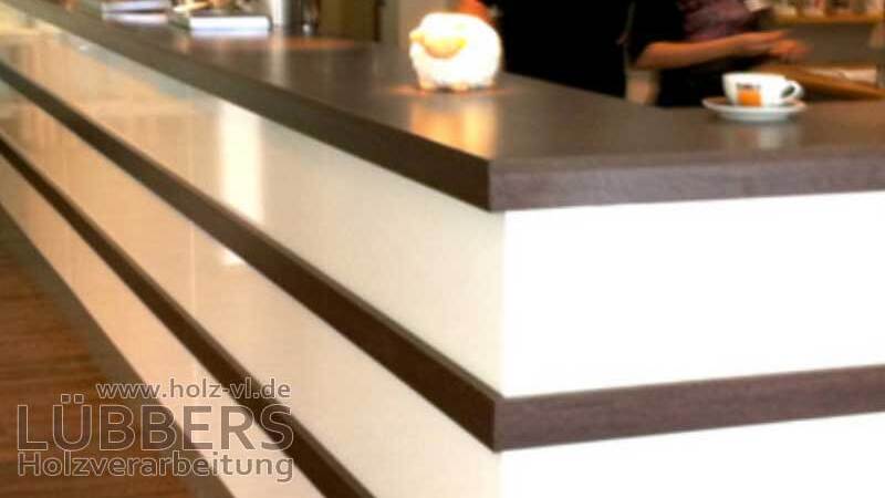 Theke mit dunkler Holzoberfl&auml;che und hellen Paneelen; im Hintergrund sind Mitarbeiter in einem Caf&eacute; aktiv.