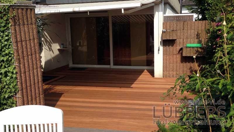 Terrasse mit Holzbelag, Blick auf gro&szlig;e Fenster des Wohnzimmers und angrenzende gr&uuml;ne Pflanzen.